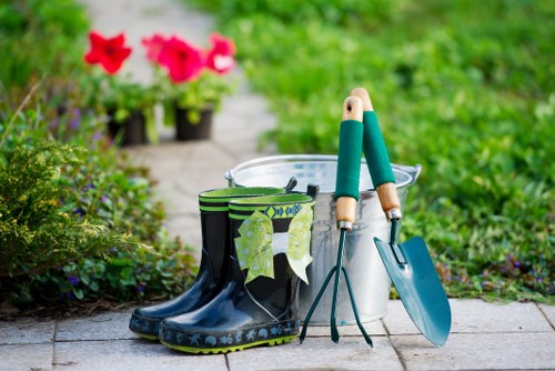 In a well-maintained front garden, a pair of black rubber gardening boots with reflective green and white design elements are placed on a paved pathway. Nearby, a pair of hand gardening tools, including a green-handled trowel and transplanter, rest inside a metallic bucket. The background showcases lush green grass and vibrant pink flowering plants in pots, indicating signs of recent gardening activity. The garden features a combination of paved walkways and natural garden beds, with a neatly edged lawn and well-kept foliage. The scene suggests a mild, possibly spring or summer day, with natural sunlight illuminating the outdoor space. This setting reflects typical gardening and lawn care elements, suitable for services provided by Hedge Trimming Barbican, emphasizing the importance of outdoor maintenance in landscaped urban gardens in Barbican or nearby areas within the postcode district.