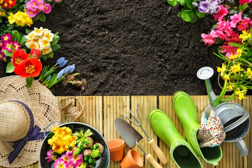 The image shows a neatly arranged outdoor garden scene with a wooden deck area bordered by a well-maintained lawn and vibrant flower beds containing a variety of colourful blooms, including pink, yellow, purple, and red flowers. On the deck, gardening tools such as a small hand trowel and a pair of green rubber gardening boots are visible, along with pots, a watering can, and a straw sunhat with a purple ribbon, indicating recent or ongoing gardening activities. The background features a section of freshly turned dark soil, suggesting preparations for planting or planting beds. The overall environment appears to be a thoughtfully landscaped garden space, likely part of a private or community outdoor area in barbican, with the scene under natural daylight, conveying a peaceful and cultivated outdoor setting suitable for gardening and outdoor maintenance services from Hedge Trimming Barbican.